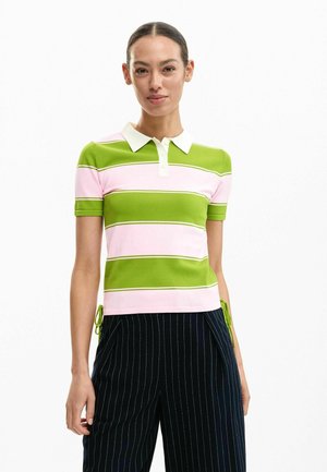 Femme portant un polo à manches courtes à rayures horizontales vertes et roses et un pantalon bleu marine à fines rayures, se tenant devant un fond blanc.