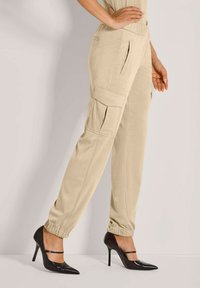 Beige Cargo-Hosen mit seitlichen Taschen und elastischen Bündchen, getragen mit schwarzen spitzen High Heels, Modell steht mit einer Hand an der Hüfte.