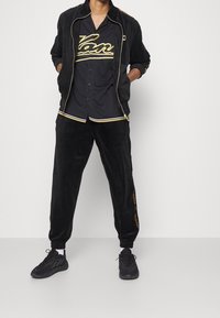 Tuta in velluto nero con accenti gialli; giacca con zip e camicia con bottoni; pantaloni abbinati con vita elastica; abbinati a sneakers nere.