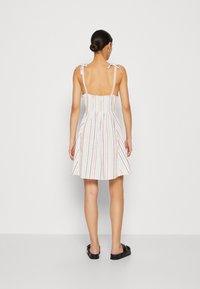 Weißes Sommerkleid mit mehrfarbigen vertikalen Streifen, Raffung am Oberteil und dünnen Trägern, die an den Schultern gebunden sind. Ausgestellter Rock und knöchellange schwarze Schuhe.