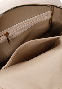 Intérieur d'un sac à main beige avec une texture de tissu lisse, présentant une poche zippée et un logo en relief. Design épuré et minimaliste.