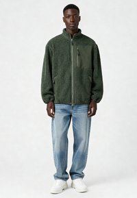 Giacca in pile verde con zip frontale e tasca sul petto, dettagli in tessuto verde abbinato, indossata con jeans azzurri e sneaker bianche.