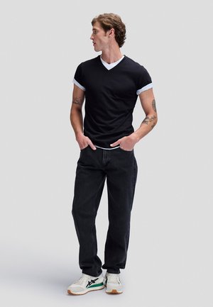 Jeune homme tatoué portant des t-shirts noirs et blancs superposés, un jean noir et des baskets blanches, debout les mains dans les poches, regardant vers la gauche.