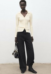Crèmekleurige geribbelde cardigan met knopen, gecombineerd met losse zwarte denim jeans. Model houdt een kleine zwarte tas vast en draagt zwarte enkellaarsjes.