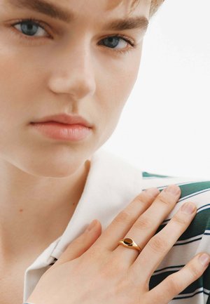 Joven con piel clara y ojos azules, vistiendo una camisa a rayas verde y blanca, con la mano en el hombro y un anillo dorado con una piedra negra en el dedo.