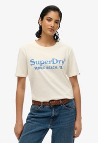Kræmfarvet bomulds t-shirt med blå grafisk tekst "SuperDry," parret med blå jeans og et brunt vævet bælte. Enkel rund halsudskæring og korte ærmer.
