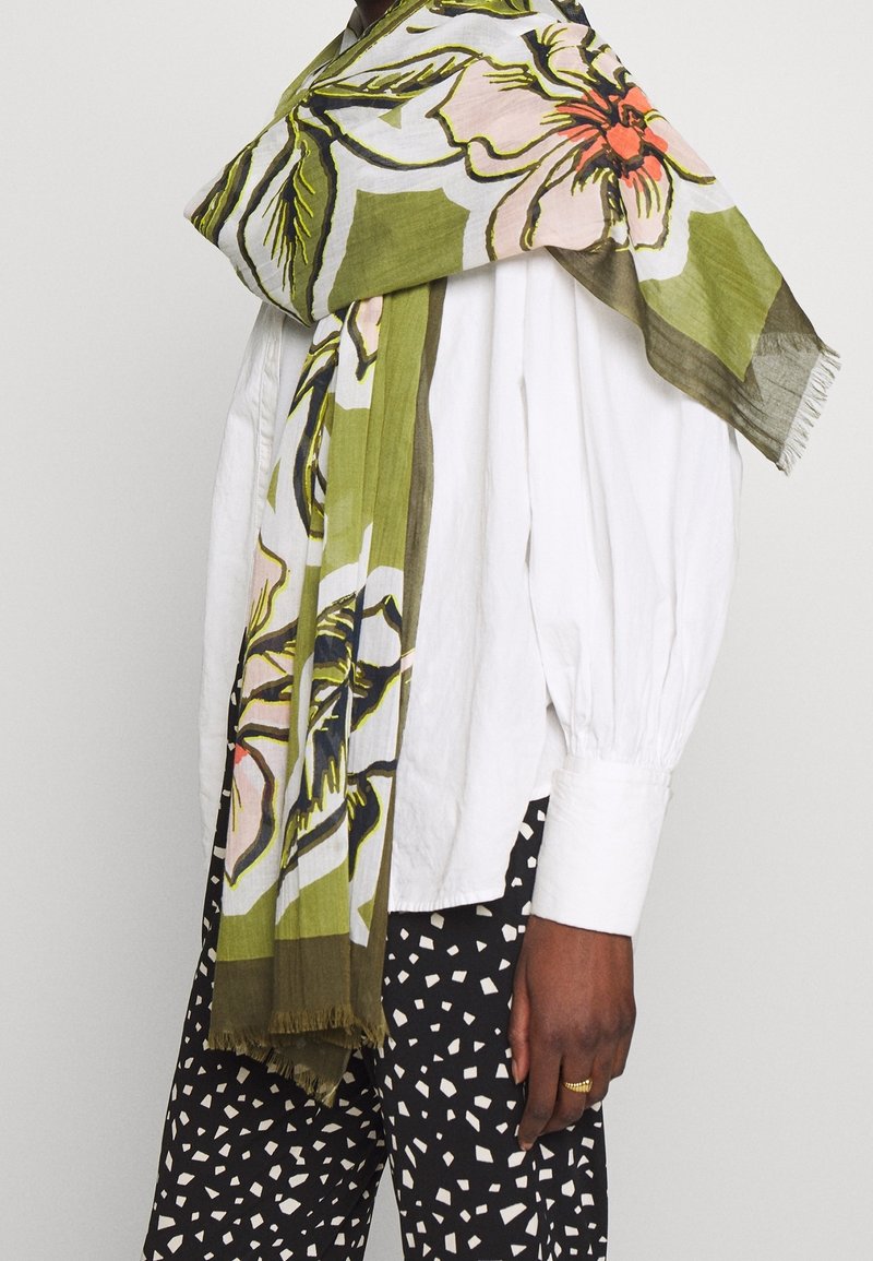 Personne portant un chemisier blanc à manches longues, un pantalon à motifs noir et blanc, et une grande écharpe verte à fleurs drapée sur les épaules.