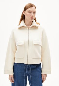 Cream-colored cropped jacket made of soft fabric, featuring a high collar, two front pockets, and a front zipper closure.