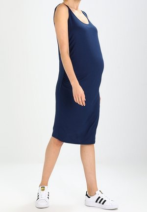 Femme enceinte debout, portant une robe bleu marine sans manches arrivant au genou et des baskets blanches avec des rayures noires.