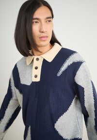 Πλεχτό πουλόβερ σε ναυτικό μπλε και γκρι με αφηρη patterns. Διαθέτει ριχτό μπεζ γιακά και τρία καφέ κουμπιά μπροστά.