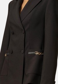 Blazer nero su misura con design doppiopetto, due tasche frontali accentuate da catena dorata e dettagli in pelle nera. Texture liscia.