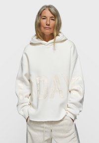Sudadera blanca con capucha con la inscripción "10 DAYS" en tela texturizada de color claro. Presenta un ajuste holgado y material suave con un diseño sencillo.
