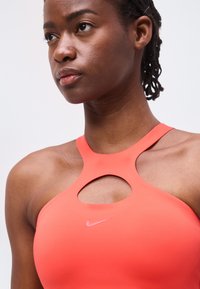 Γυναίκα με κοραλί αθλητικό τοπ χωρίς μανίκια με άνοιγμα σε σχήμα κλειδιού και λογότυπο Nike, φαίνεται συγκεντρωμένη σε απλό φωτεινό φόντο.