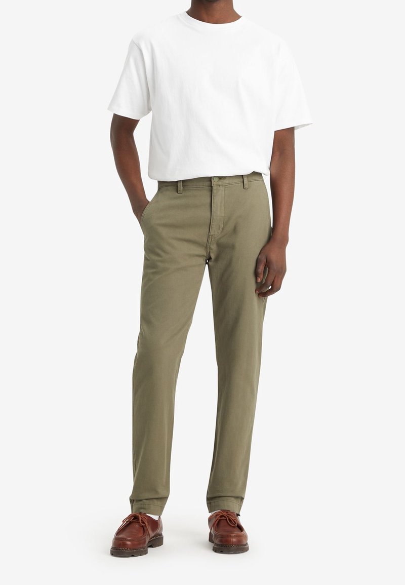 Olivgrüne Chinos mit geradem Schnitt, Baumwollstoff, Vordertaschen und Gürtelschlaufen, kombiniert mit einem schlichten weißen T-Shirt und braunen Schuhen.