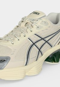 Μπεζ και ασημί παπούτσι για τρέξιμο Asics με επάνω μέρος από δίχτυ, μαύρες και ασημί λωρίδες με λογότυπο, λευκά σχέδια στα κορδόνια και πράσινο μαξιλάρι στη σόλα.