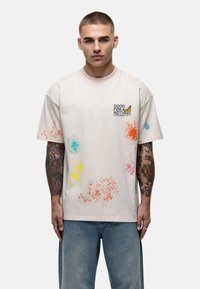 Biała bawełniana koszulka z grafiką motyla i napisem "GOOD FOR NOTHING", ozdobiona plamami niebieskiego, żółtego, różowego i pomarańczowego.