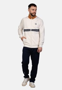 Weiße Zip-Jacke mit marineblauen Streifenakzenten, getragen über einem braunen hochgeschlossenen Oberteil, kombiniert mit navyfarbenen Hosen und beigen Sneakern. Legere Bekleidung.