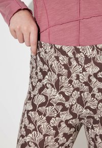 Maglietta a maniche lunghe color rosa abbinata a pantaloni marroni con fantasia, caratterizzati da disegni fogliari bianco sporco, che evidenziano la texture e la vestibilità del tessuto.