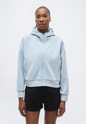 Sieviete stāv, ar seju pret priekšu, valkājot gaiši zilu rāvējslēdzēja kapuci un melnas šortus, vienkrāsainas baltas fona priekšā.