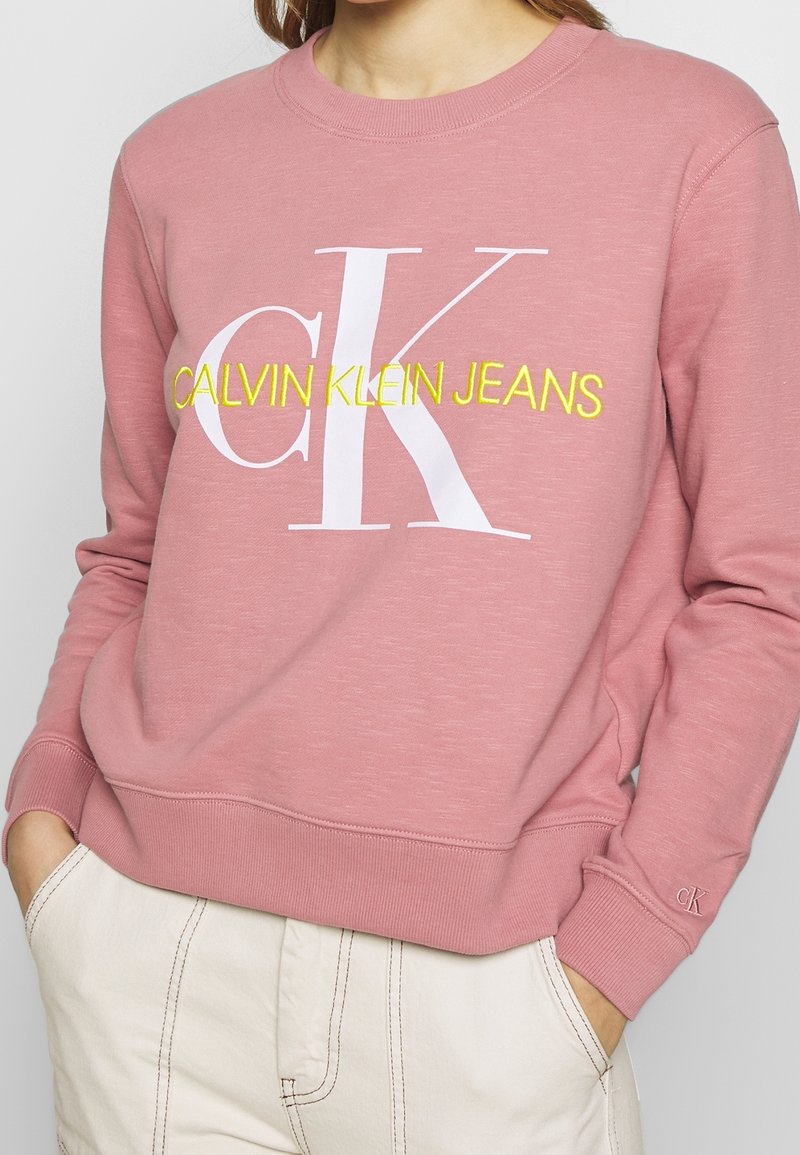 Sudadera de algodón rosa con puños y dobladillo acanalados, que presenta un gran logo blanco "CK" y el texto amarillo "CALVIN KLEIN JEANS". Bolsillo frontal visible.