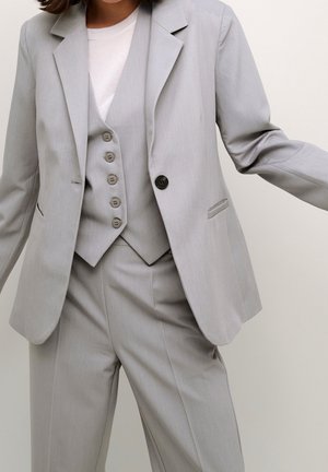 Kobieta w jasnoszarym trzyczęściowym garniturze z zapinanym na guziki kamizelką, marynarką i spodniami, ramiona lekko rozszerzone na jednolitym tle.