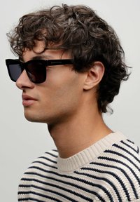 Sunglasses med svarta glasögonbågar och mörka linser, med en modern fyrkantig form. Bärs av en person i en randig krämfärgad och marinblå tröja.