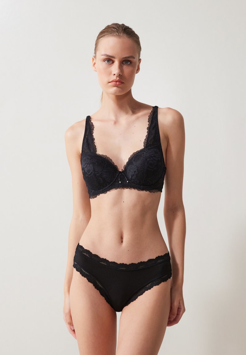 Μαύρο δαντελένιο σουτιέν με φαρδιές τιράντες και κομμένο τελείωμα, συνδυασμένο με αντίστοιχες μαύρες εσώρουχες με δαντελένιο φινίρισμα, που διαθέτουν λεία υφή.