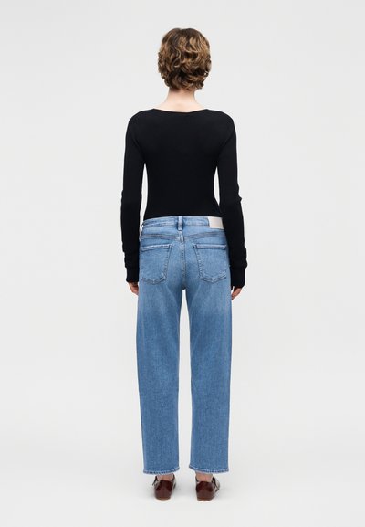 Γυναίκα με κοντά σγουρά μαλλιά, φορώντας εφαρμοστή μαύρη μπλούζα με μακριά μανίκια, τζιν παντελόνι ευθύγραμμης γραμμής σε ανοιχτό μπλε και καφέ παπούτσια, στέκεται γυρισμένη προς τα πίσω.