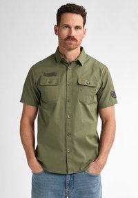 Camisa de manga corta de algodón verde con botones, con dos bolsillos en el pecho y un parche en la manga, que presenta una textura suave y un corte suelto.