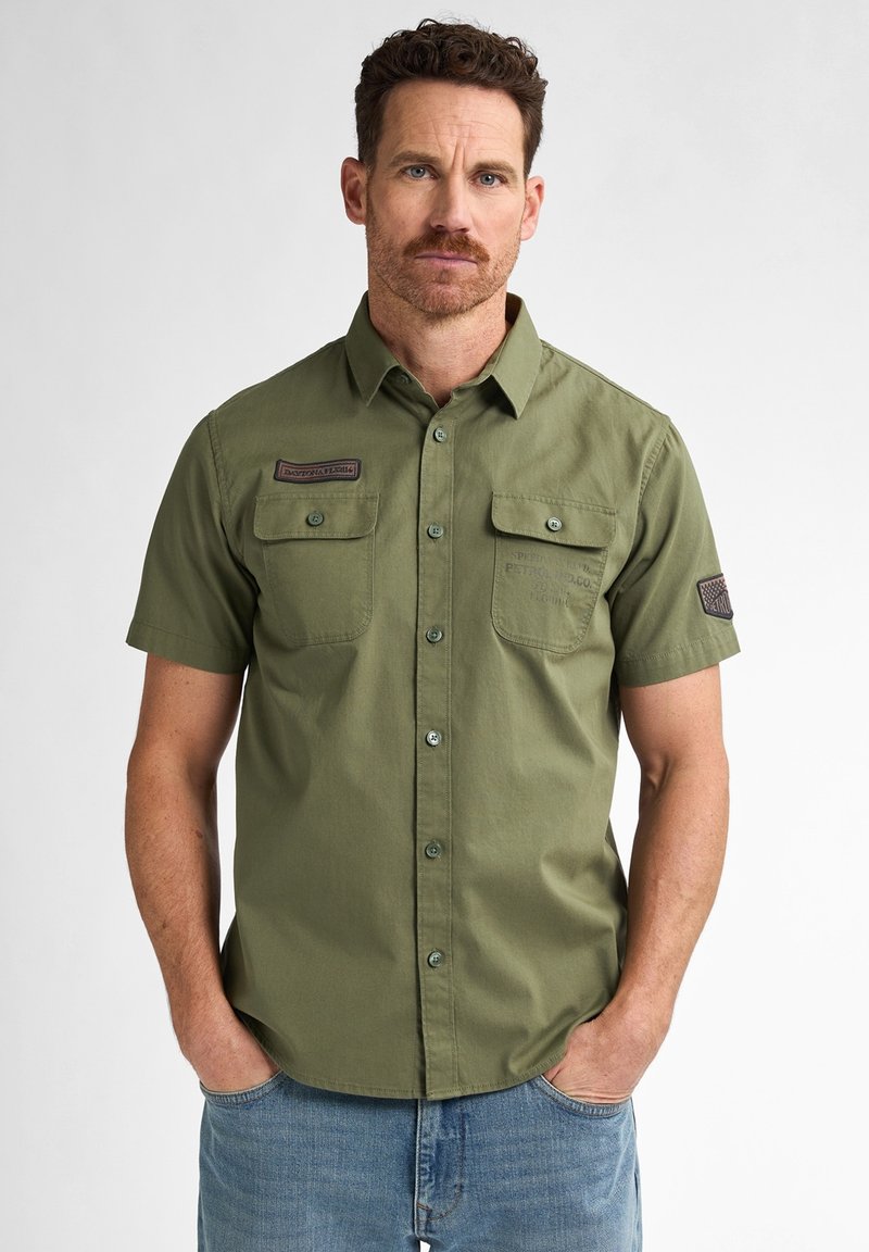 Camisa de manga corta de algodón verde con botones, con dos bolsillos en el pecho y un parche en la manga, que presenta una textura suave y un corte suelto.