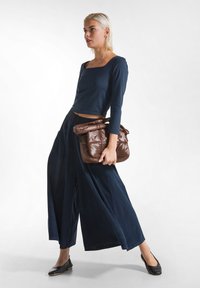 Marineblauwe top met lange mouwen en wijde broek van soepel materiaal, gecombineerd met een bruine puffer tas en zwarte platte schoenen, in een minimalistische omgeving.