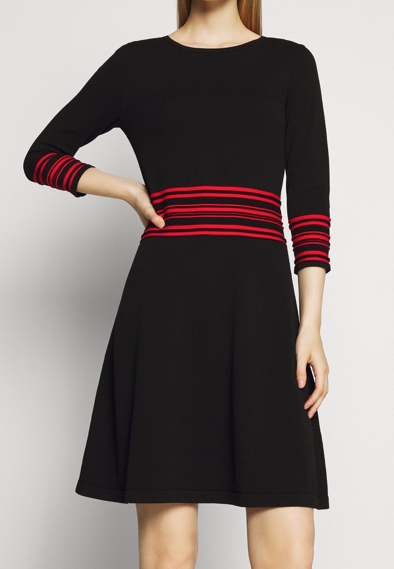 Femme portant une robe noire à manches trois-quarts avec des rayures rouges horizontales à la taille et aux poignets des manches.