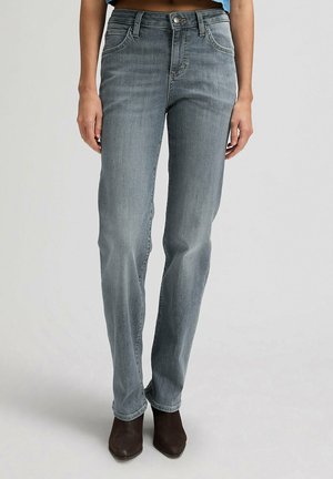 Kobieta ubrana w wyblakłe szare dżinsy o prostych nogawkach i ciemnobrązowe botki z noskiem w szpic, stojąca na tle jednolitego jasnego tła.