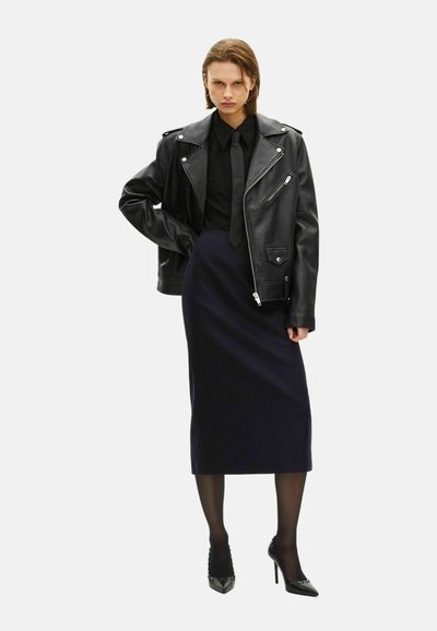 Giacca moto in pelle nera sopra una camicia e cravatta nera, abbinata a una gonna midi blu navy e tacchi alti neri, creando una silhouette aderente.