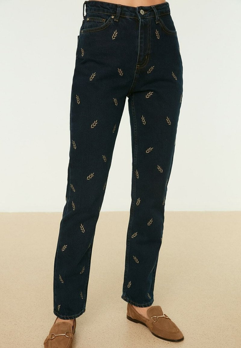 Jean en denim bleu foncé avec petits motifs de feuilles beige, porté avec des mocassins marron sur un sol beige.