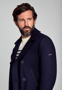 Cappotto di lana blu navy con design doppiopetto, grandi bottoni e rifinitura a contrasto sulle tasche, indossato sopra un maglione a righe color crema.