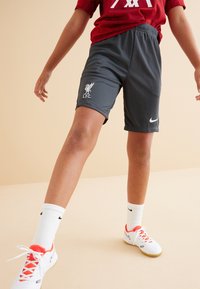 Graue Sportshorts mit elastischem Bund und weißem Liverpool FC Logo. Das Model trägt weiße Socken und Nike-Schuhe mit orangefarbenen Akzenten.