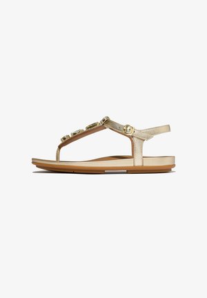 Gold flat sandal with ankle strap, buckle closure, and decorative gems on the T-strap, shown in side profile against white background.