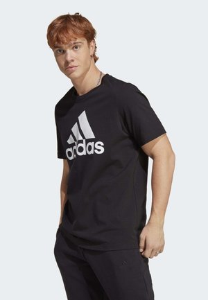Zwarte katoenen t-shirt met een grote witte Adidas-logo op de voorkant. Korte mouwen en een ontspannen pasvorm, gecombineerd met zwarte broek.