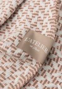 Tkany tekstyl, z różowo-beżowym wzorem, z satynową metką z napisem "L'ATELIER de Paris." Miękka faktura, subtelny blask.