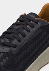 Gros plan sur une basket en cuir noir tressé avec des lacets noirs, une doublure intérieure beige et une semelle blanche texturée.