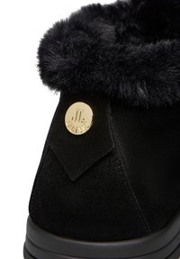 Schwarze Wildlederstiefel mit einem Kragen aus Kunstfell, flacher schwarzer Sohle und einem runden goldenen Logo-Akzent an der Seite. Texturierte Oberfläche auf dem Material sichtbar.