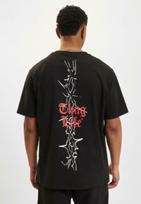 Čierne bavlnené tričko s krátkymi rukávmi, ktoré na prednej strane zobrazuje vertikálny biely grafický prvok s nápisom "Thug Life" v červenom texte a na zadnej strane má dizajn pripomínajúci tŕne.