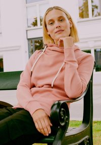Rózsaszín kapucnis pulóver, cseppnyakú kivitelben, fehér húzózsinórokkal. Puha tapintás; a modellen fekete nadrág van, ül egy zöld padon, ellazult testtartásban.
