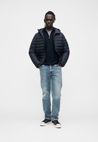 Μπουφάν puffer σε σκούρο μπλε, φορεμένο πάνω από μπλε ζακέτα ναυτικού τύπου, σετ με ανοιχτό μπλε τζιν με φθορές και μαύρα αθλητικά παπούτσια με λευκές σόλες.