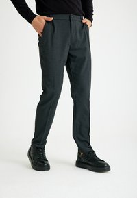 Graue maßgeschneiderte Hosen mit schlanker Passform, ausgestattet mit einem Knopfverschluss, seitlichen Taschen und glatter Oberfläche. Kombiniert mit schwarzen Sneakers.