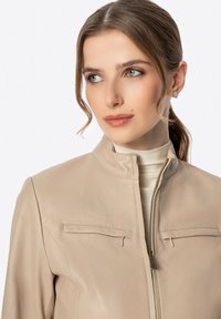 Veste en cuir beige avec un devant zippé, deux poches poitrine, col montant et texture lisse. Le modèle porte un haut clair en dessous.