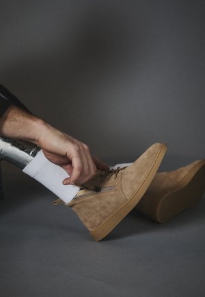 Main gauche ajustant la fermeture éclair d'une botte cheville en daim beige portée sur une chaussette blanche sur une jambe prothétique métallique sur fond gris.