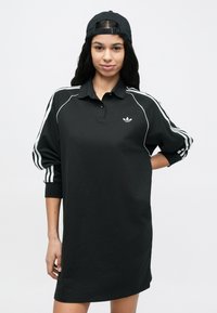 Μαύρο φόρεμα πόλο από βαμβάκι, με λευκές ρίγες στα πλάγια και μικρό λογότυπο στο στήθος. Χαλαρή εφαρμογή, μακριά μανίκια και γιακάς.