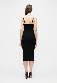 Γυναίκα με μακριά ίσια μαλλιά, που φοράει μαύρο μίντι φόρεμα με αλυσίδες στους ιμάντες και μαύρα παπούτσια με ψηλό τακούνι, γυρισμένη προς τα πίσω, σε απλό φόντο.